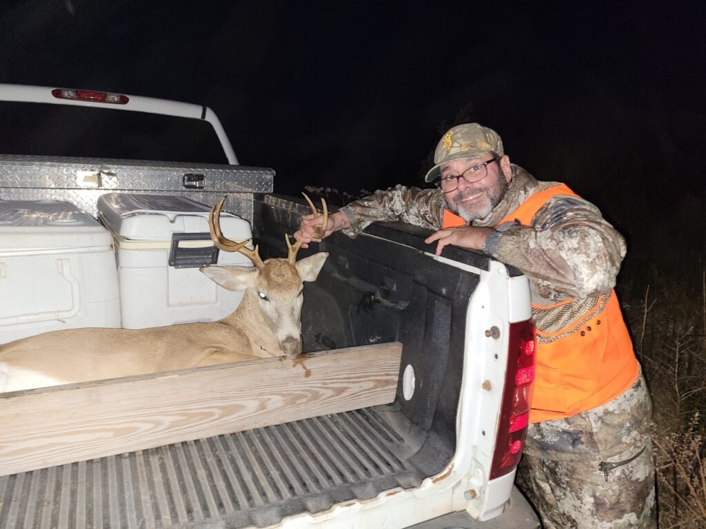 Hunter Wearing Camouflage and an Orange Vest Smiling Beside a Harvested Deer Loaded in the Back of a Pickup Truck at Night