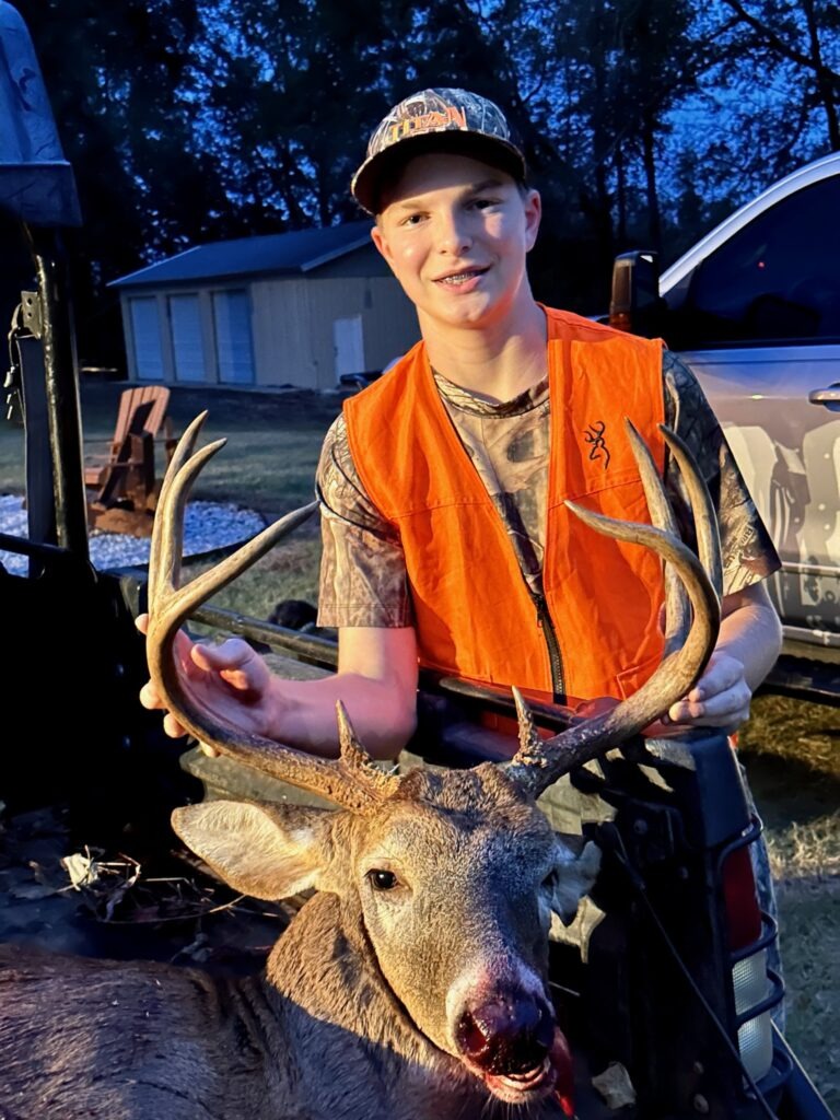 Young Hunter Wearing an Orange Safety Vest Posing with a Harvested Deer During Dusk Near a Pickup Truck