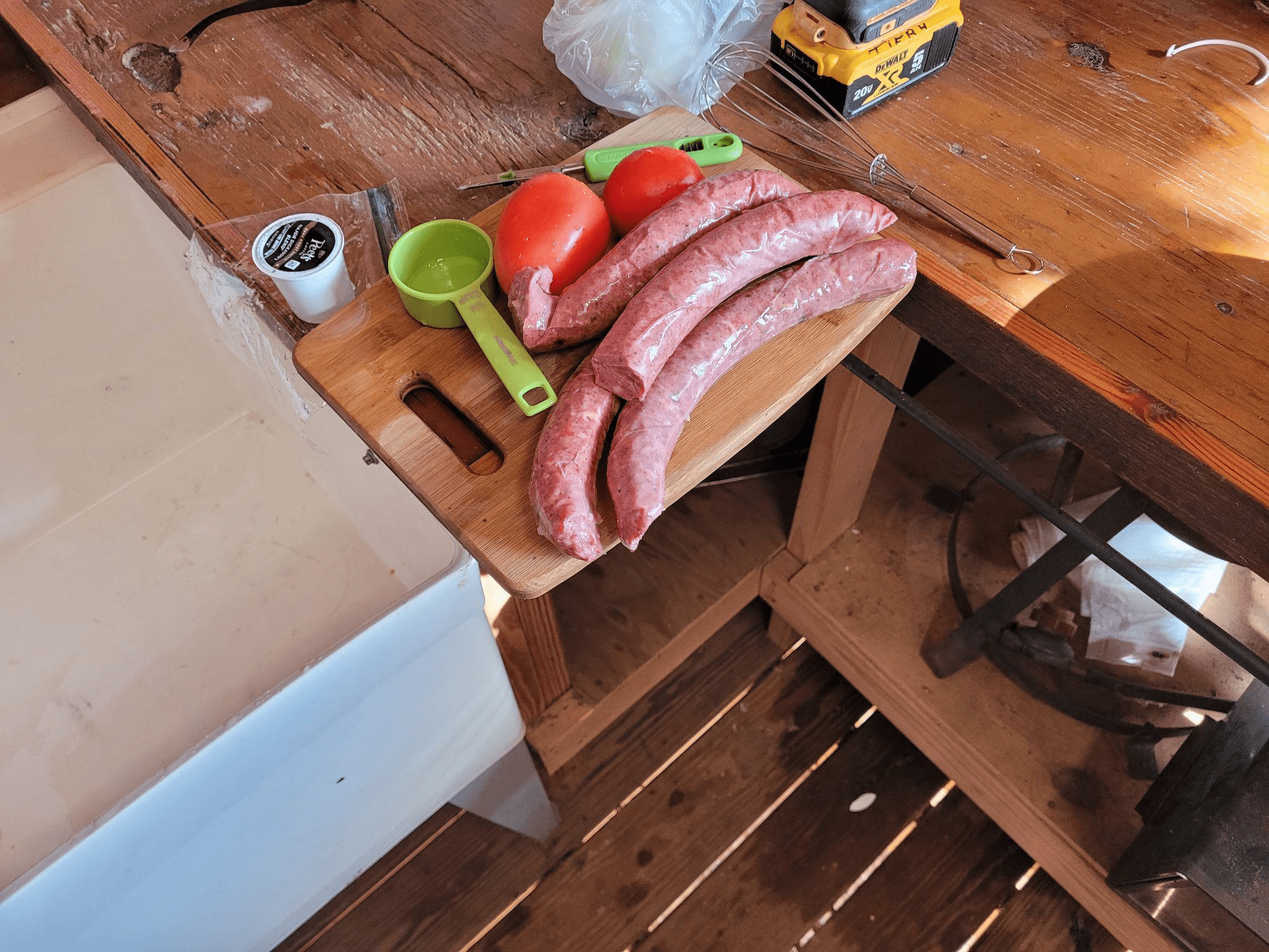 Sliced sausages, tomatoes, and measuring cups are arranged on a wooden cutting board, indicating preparation for cooking in a rustic kitchen.